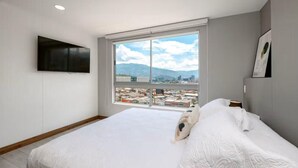 Un dormitorio con una ventana grande, una cama, un televisor de pantalla plana y vistas a la ciudad.