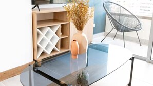 Una sala de estar moderna con una mesa de centro de vidrio, un mueble de madera con estantes, un jarrón con plantas secas y una silla de alambre negra.