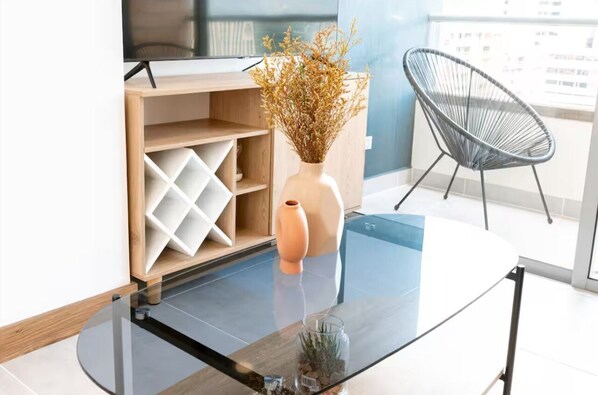 Una sala de estar moderna con una mesa de centro de vidrio, un mueble de madera con estantes, un jarrón con plantas secas y una silla de alambre negra.