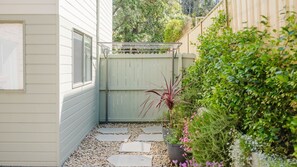 A narrow pathway with stepping stones leading to a wooden gate, surrounded by lush greenery and a white building.