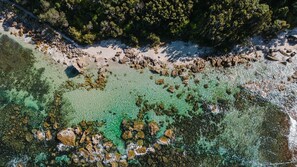 An aerial view of a beach with clear turquoise waters, rocky formations, and dense forested areas.