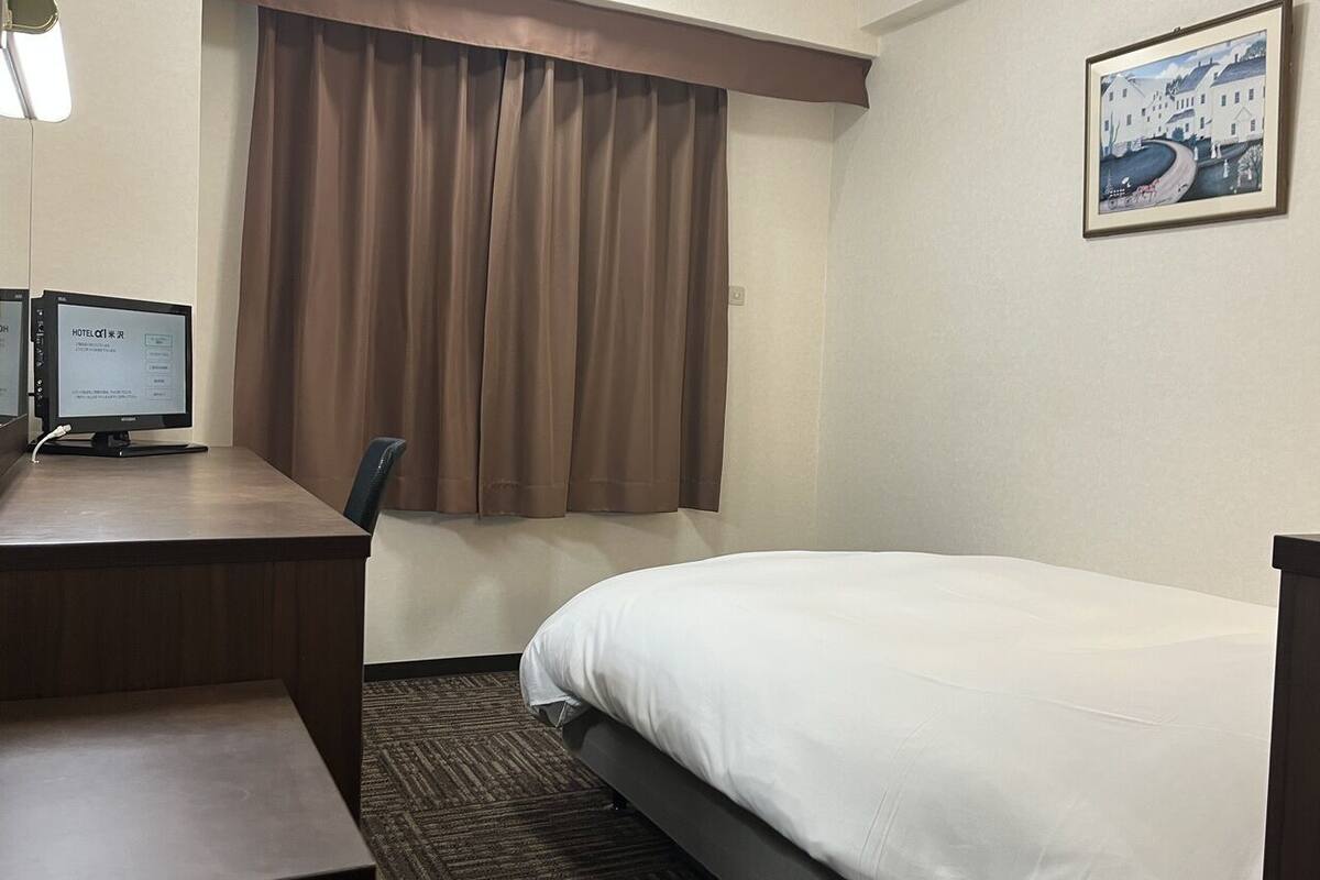 A hotel room with a bed, a desk with a computer, a chair, a window with curtains, and a framed picture on the wall.