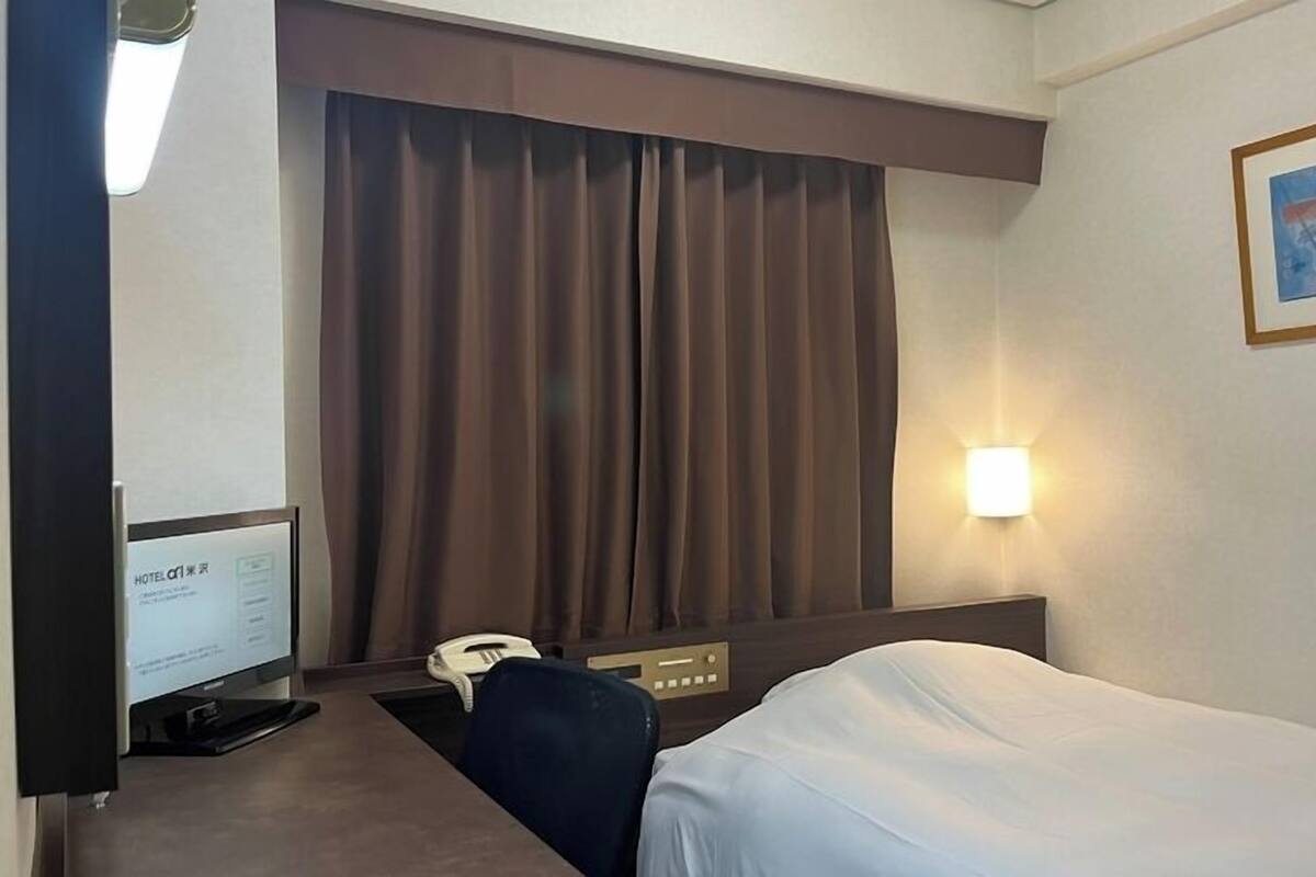 A hotel room with a bed, a desk with a computer and phone, a large window with curtains, and a wall-mounted light.