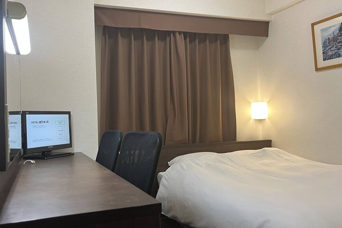 A hotel room with a bed, a desk with a computer, two chairs, a window with curtains, and a wall-mounted light.
