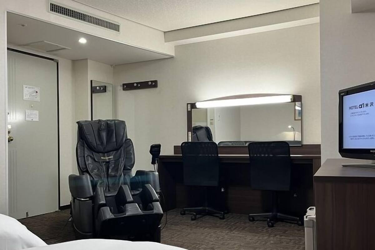 A hotel room with a bed, a massage chair, a desk with a computer, and a large mirror.