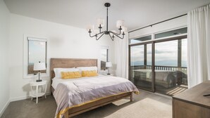 Un dormitorio con cama de madera, ropa de cama blanca, una araña, una mesita de noche pequeña y una cómoda, además de un gran ventanal corredero que da a un balcón con vistas a las montañas.