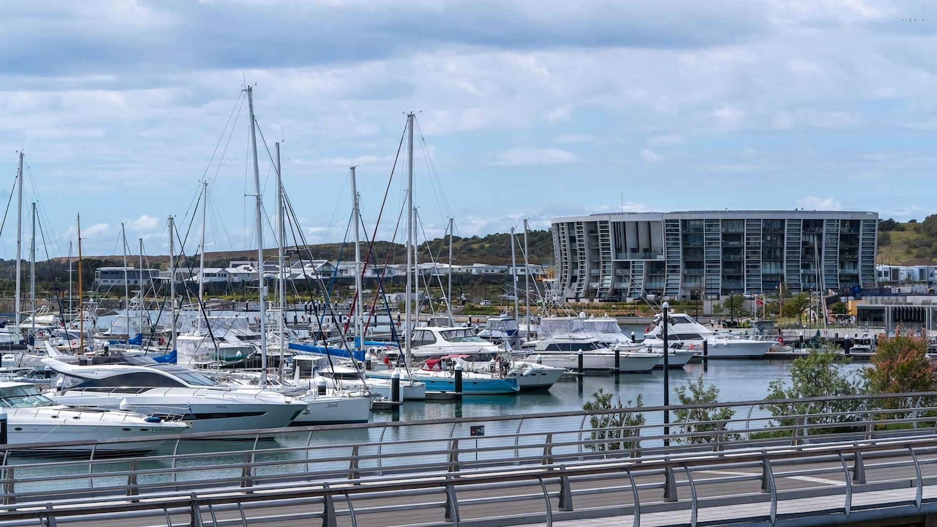 Un port de plaisance où sont amarrés de nombreux bateaux, un bâtiment moderne en arrière-plan et un ciel dégagé.