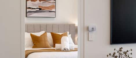 Una cama bien hecha con ropa de cama blanca y marrón, un cabecero gris y un termostato fijado a la pared.
