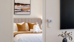Una cama bien hecha con ropa de cama blanca y marrón, un cabecero gris y un termostato fijado a la pared.