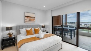 Un dormitorio moderno con una cama grande, una mesita de noche, una lámpara de noche y vistas a un balcón con mobiliario exterior.