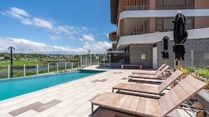 Un espace aménagé au bord de la piscine, avec des chaises longues et des parasols, un ciel dégagé et une vue sur un quartier résidentiel.
