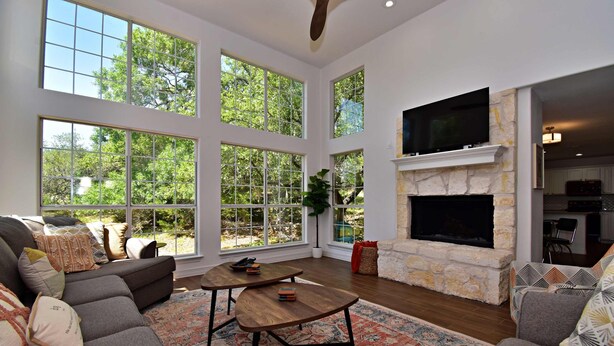 A spacious living room with a stone fireplace, large windows, and a cozy seating area. - Dreamy Dripping Springs | Near Parks, Hiking & More! (Dripping Springs)