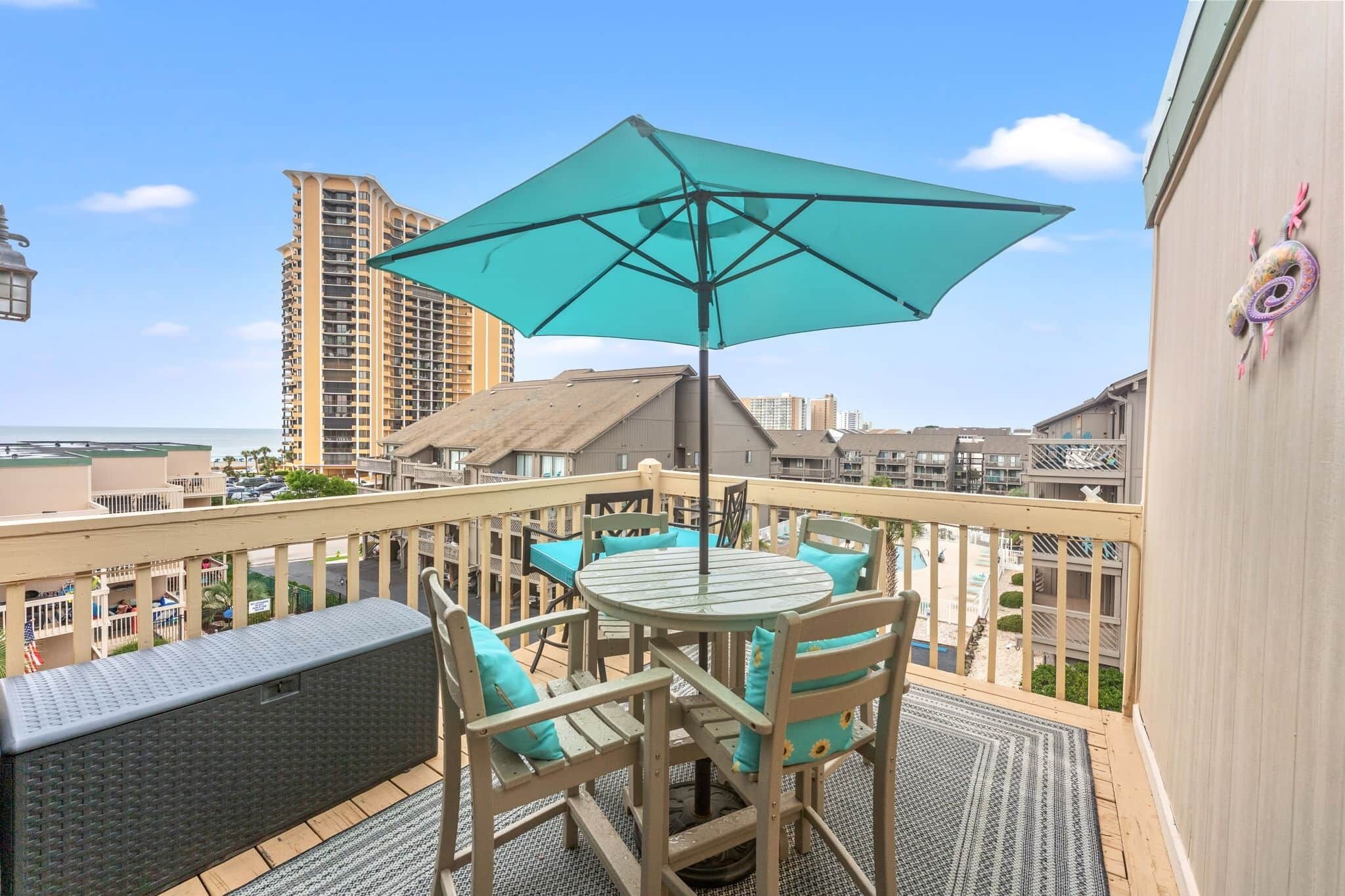 A balcony with a table and chairs, a turquoise umbrella, and a view of a tall building and a pool.