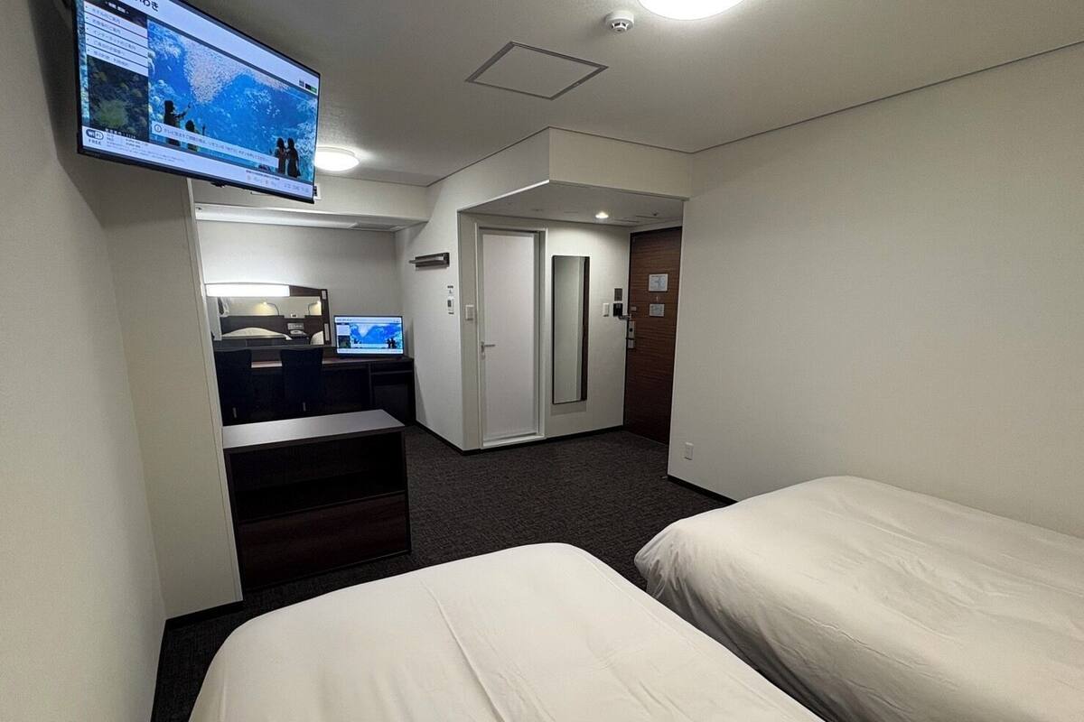 A hotel room with two beds, a desk with a computer, a television mounted on the wall, and a door leading to another room.