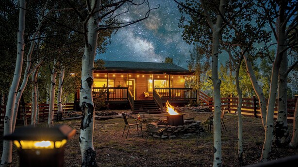 Una cabaña con una fogata y asientos exteriores, rodeada de árboles y un cielo nocturno estrellado.