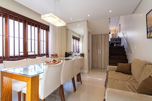 Una sala de estar moderna con una mesa de comedor de vidrio, sillas blancas, un sofá marrón y una escalera que sube a un nivel superior.