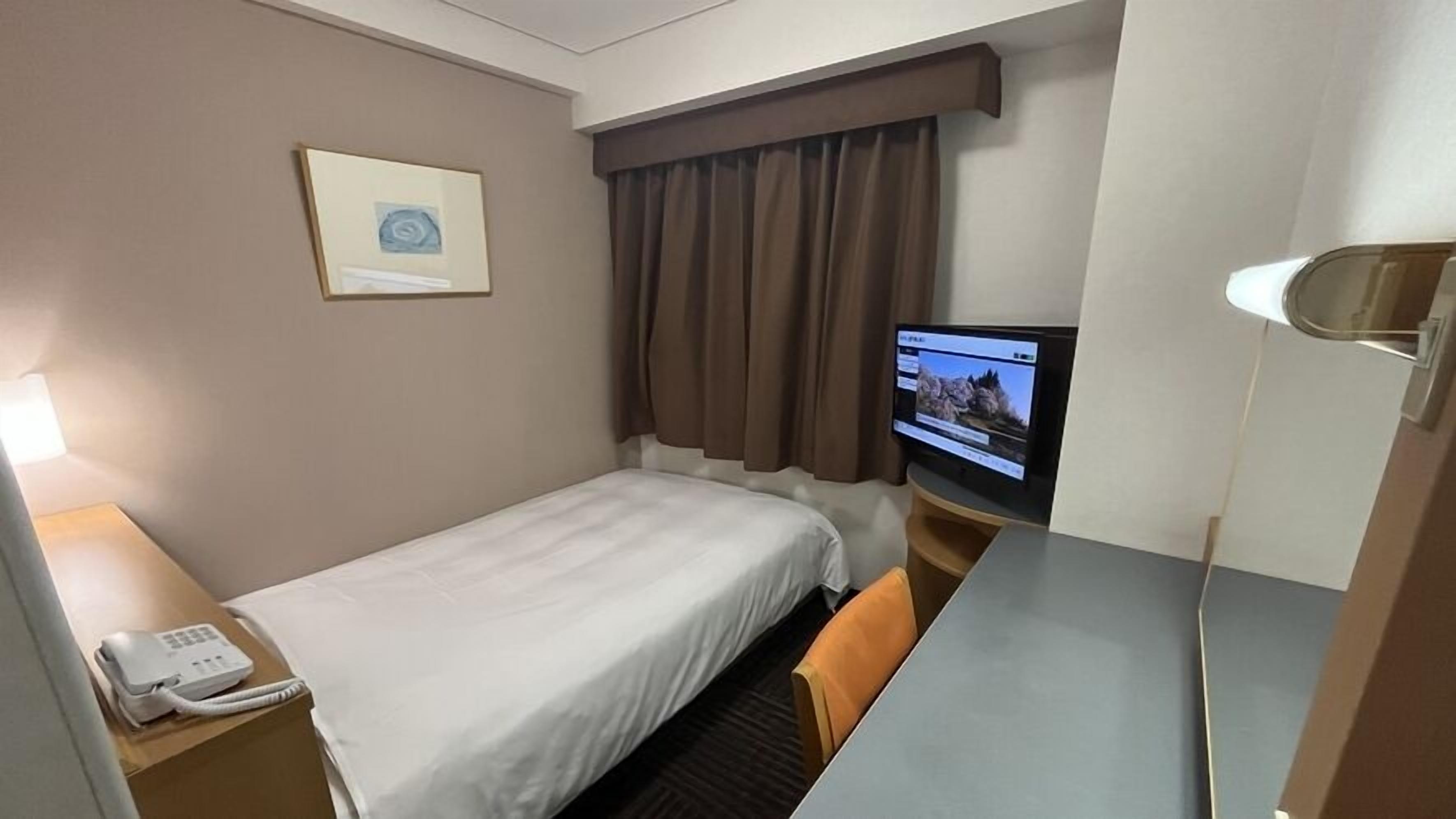 A hotel room with a single bed, a desk with a computer monitor, a telephone, and a window with curtains.