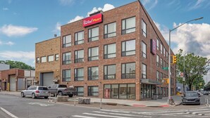A multi-story brick building with a Red Roof Plus+ sign, large windows, and a street view with cars and a crosswalk. - Red Roof Inn PLUS+ Brooklyn - 3rd Ave (Brooklyn)