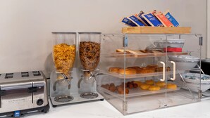 Estação de pequeno-almoço de serviço livre, com distribuidores de cereais, pão, pastelaria e saquinhos de café.