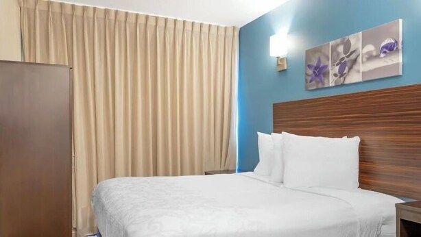 Quarto de hotel com cama, cabeceira de madeira, cortinas bege e parede com pintura floral.