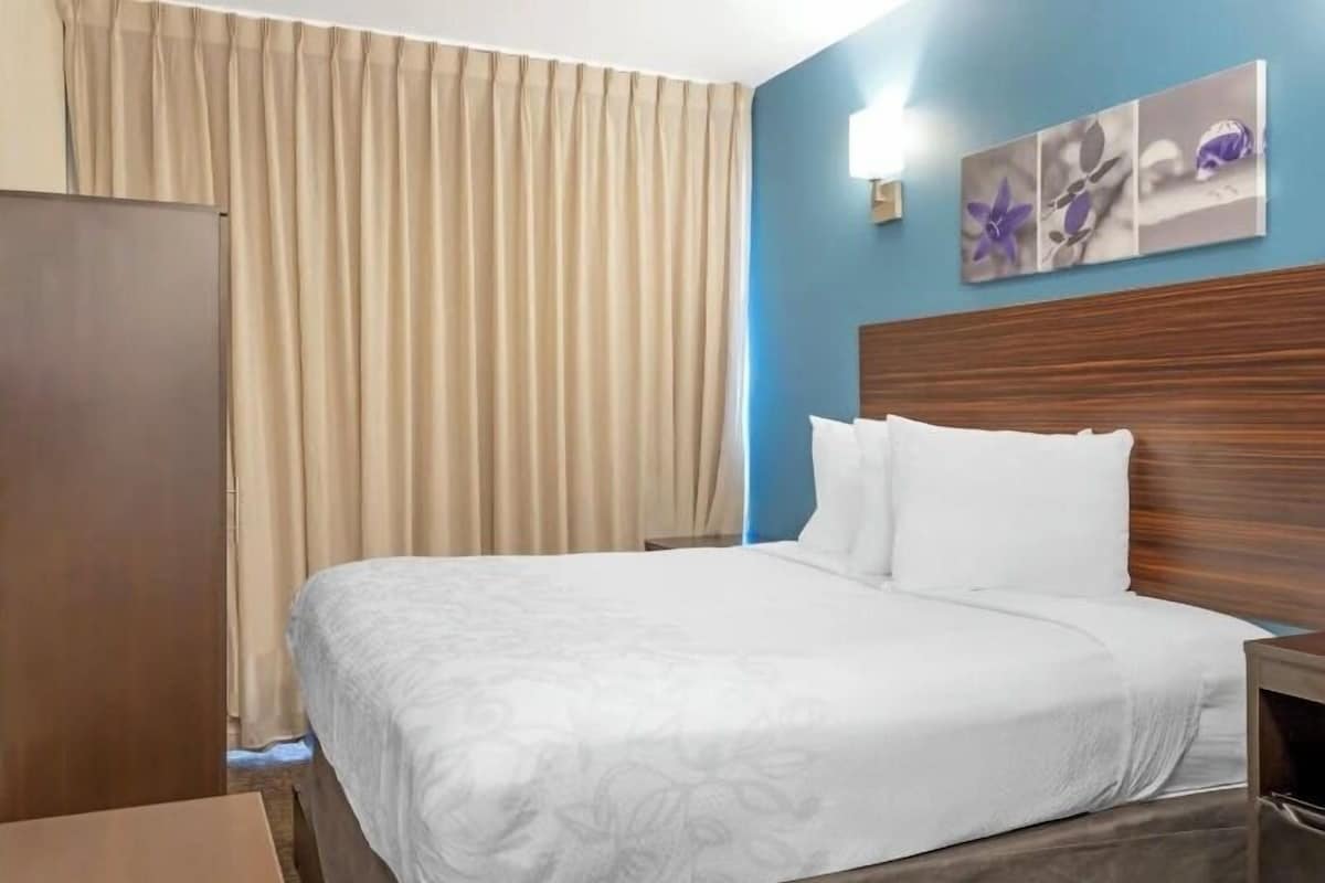 Habitación de hotel con una cama, cabecera de madera, cortinas color beige y un cuadro floral en la pared.