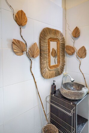 Une salle de bain avec un lavabo en pierre, un miroir doté d’un cadre tressé et des éléments décoratifs muraux représentant des feuilles et des branches.