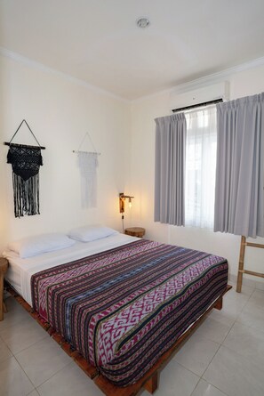Une chambre avec un lit, un climatiseur fixé au mur, une décoration murale en macramé, une lampe de chevet et une fenêtre avec des rideaux.