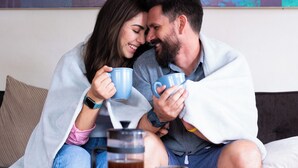 Un couple est assis sur un canapé, une table basse se trouve devant eux, et ils tiennent chacun une tasse.