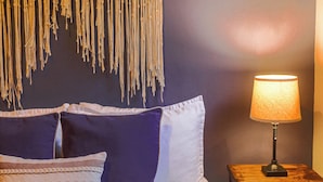 Un lit bien fait, avec une couvre-lit à motifs et des oreillers, une décoration murale en macramé, une lampe de chevet et une petite table de nuit en bois sur laquelle est pliée une serviette.