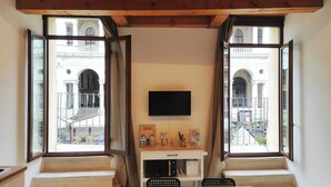 Une pièce avec un téléviseur fixé au mur, un bureau avec un ordinateur et un lit. Il y a deux fenêtres donnant sur un bâtiment orné d’arcades.