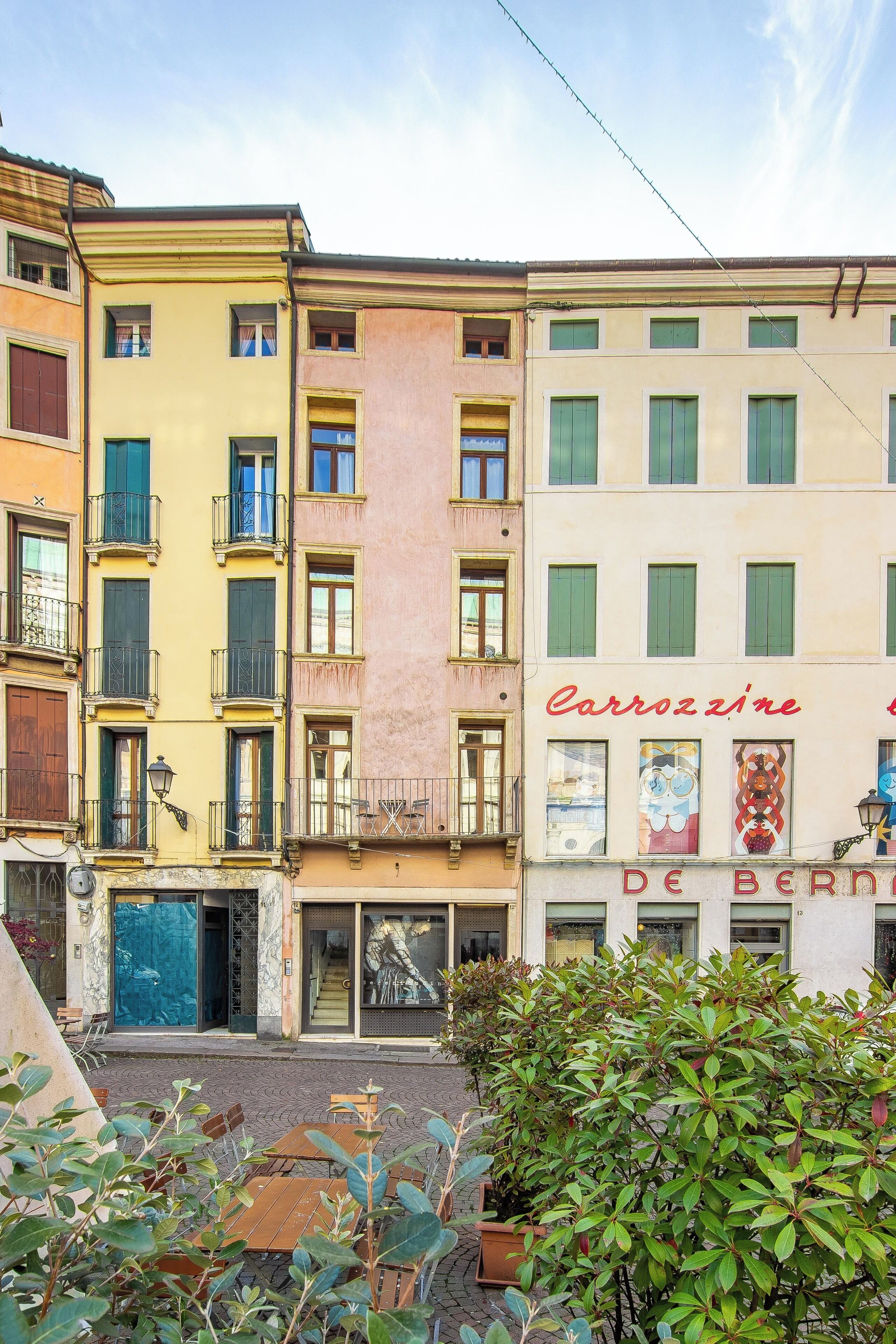Une rangée d’immeubles à plusieurs étages dotés de balcons, une rue devant, et un petit espace jardin avec des plantes en pot.