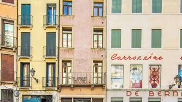 Une rangée d’immeubles à plusieurs étages dotés de balcons, une rue devant, et un petit espace jardin avec des plantes en pot.