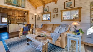 Ein gemütliches Blockhaus-Interieur mit Holzbalkendecke, einem Wohnzimmer mit einer Ledersofa, einem Couchtisch und einer Küchenzeile inklusive Herd und Regalen.