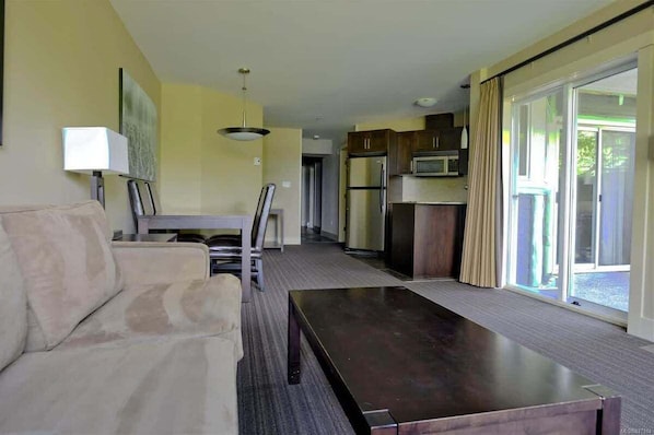 A modern living room with a sofa, a coffee table, and a dining area with a table and chairs. There is a kitchen area with cabinets and appliances.