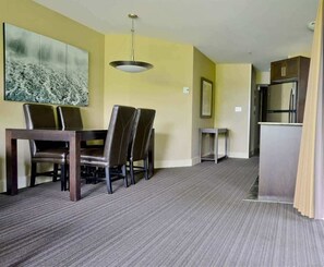 A hotel room with a dining area featuring a table and chairs, a small side table, a ceiling light, and a kitchenette with a refrigerator and cabinets.