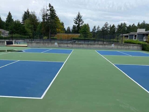 An outdoor tennis court with multiple courts, surrounded by trees and a fence.