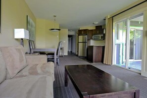 A modern living room with a sofa, a coffee table, and a dining area with a table and chairs. There is a kitchen area with cabinets and appliances.