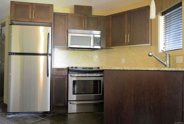 Una cocina con armarios de madera oscura, electrodomésticos de acero inoxidable, incluyendo un refrigerador, microondas y horno, y una encimera de granito.