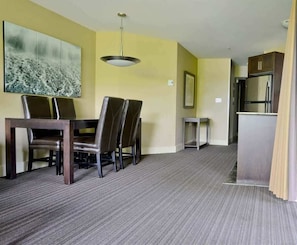 A hotel room with a dining area featuring a table and chairs, a small side table, a ceiling light, and a kitchenette with a refrigerator and cabinets.