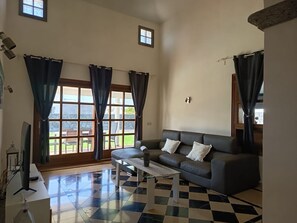 A living room with a sofa, a coffee table, and a TV. There are curtains on the windows, and a view of the outside. - Beautiful stylish villa with 2 bedrooms, private pool and Jacuzzi. (Playa Blanca)