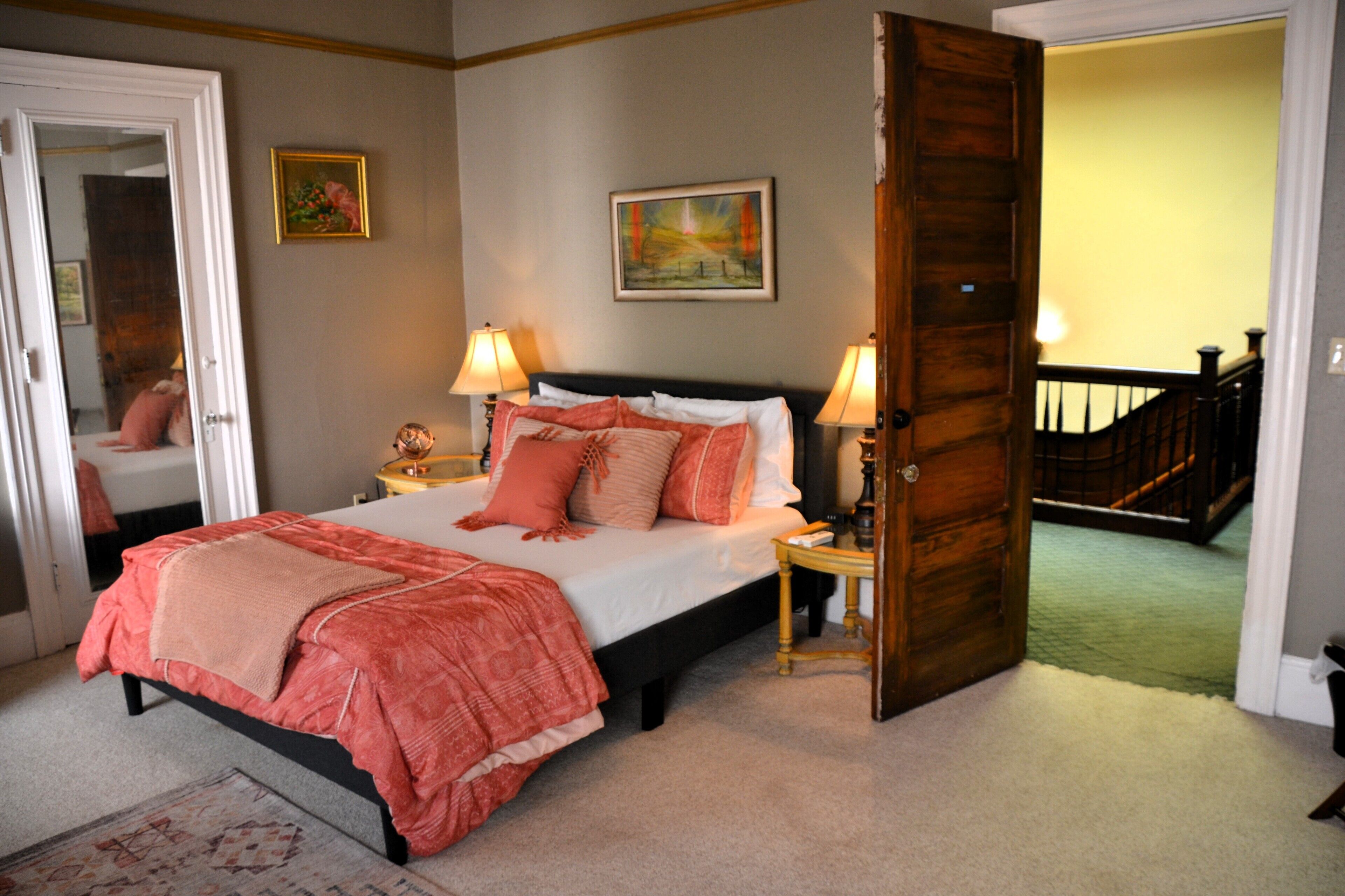 A bedroom with a bed, two bedside tables, a door, a painting, and a staircase.