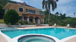 Uma casa de dois andares com piscina, banheira de hidromassagem e área de estar no pátio com espreguiçadeiras.