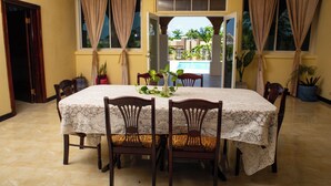 Uma área de refeições com uma mesa coberta por um tablecloth estampado, rodeada por cadeiras de madeira com assentos acolchoados. Há plantas em vasos sobre a mesa e ao longo das paredes.