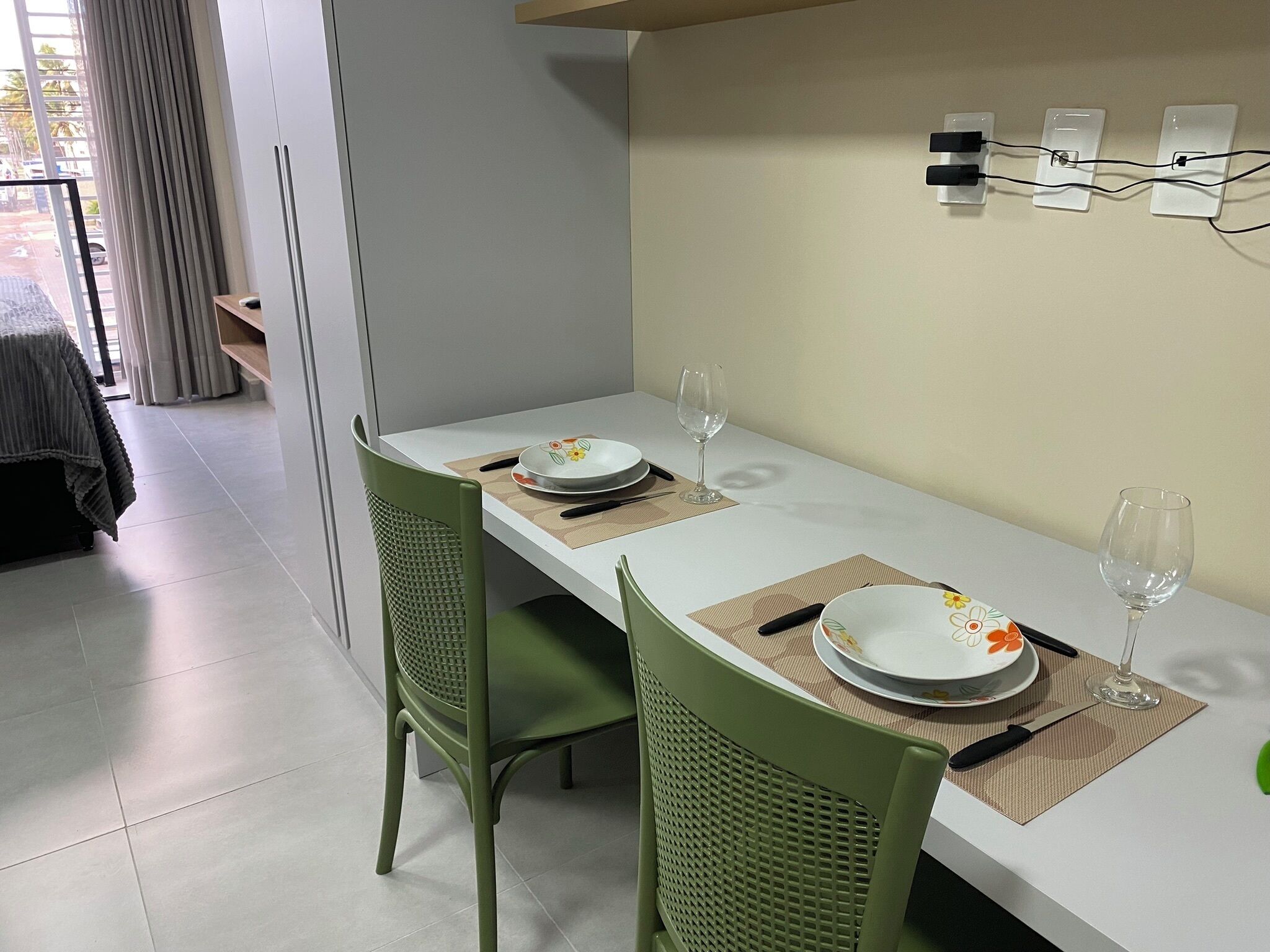Un coin repas avec une table blanche, deux chaises vertes et un luminaire fixé au mur.