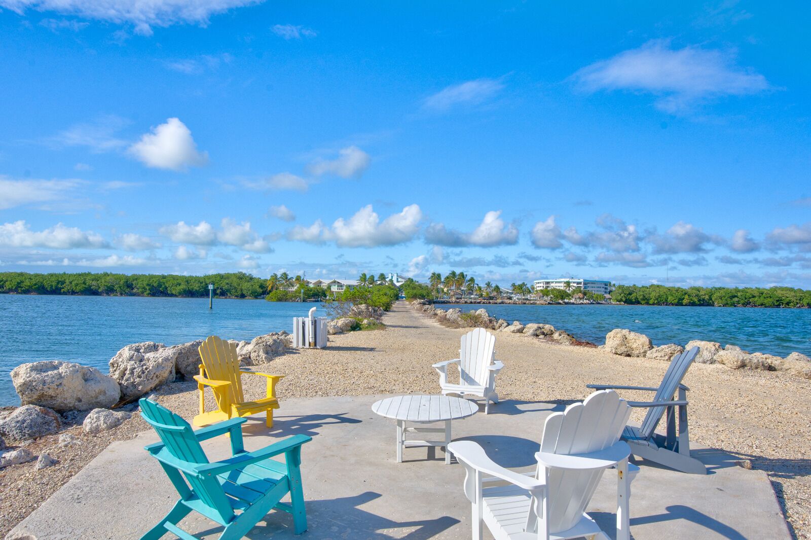 A beachside seating area with Adirondack chairs and a table, overlooking a calm body of water with a distant shoreline and buildings.