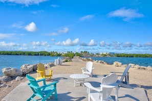 Uma área de estar à beira-mar com cadeiras Adirondack e uma mesa, com vista para águas calmas e uma linha de costa distante com edifícios.