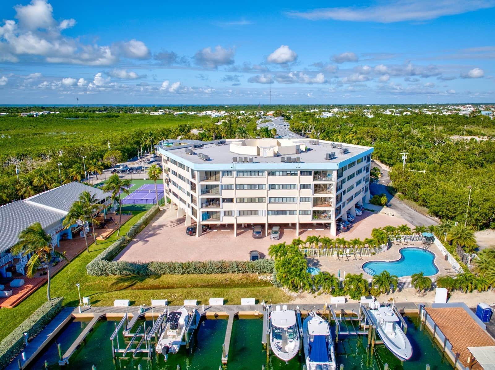A multi-story hotel with a rooftop pool, surrounded by greenery and a marina with boats.