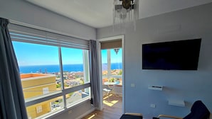 Habitación de hotel moderna con un amplio ventanal que ofrece vistas a la ciudad y al océano, una cama, un televisor y un pequeño rincón de estar.