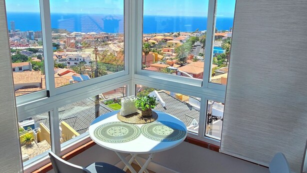 Un balcón con una mesa, dos sillas y vistas a un pueblo costero y al mar.