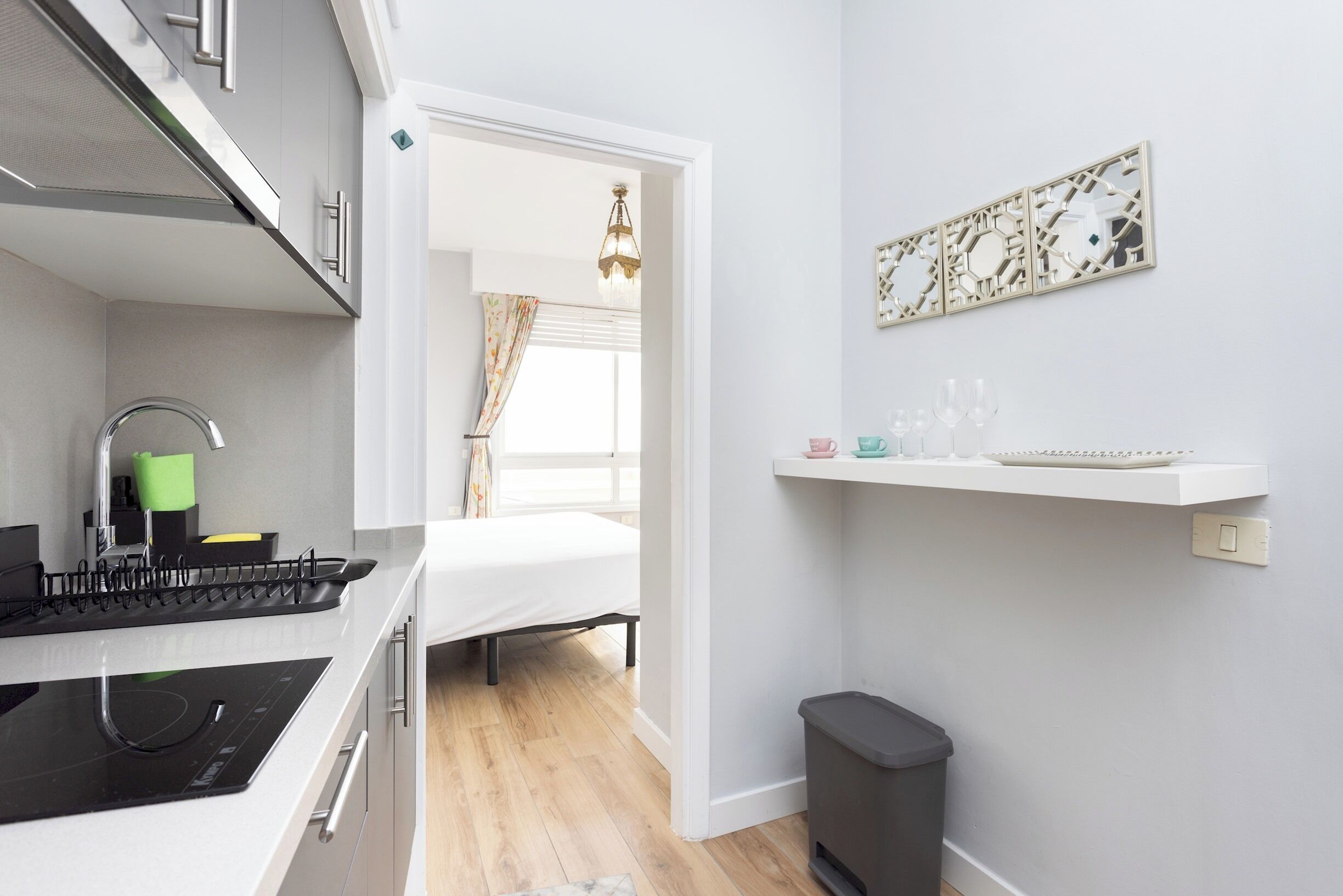 A compact kitchen with a stove, sink, and a small trash bin. A bed is visible through an open doorway, and a decorative wall piece hangs above a shelf with glasses.